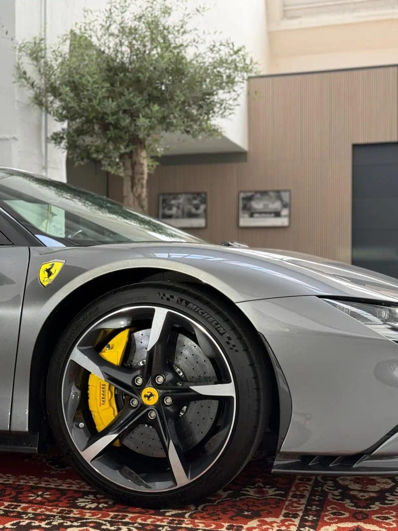 Nahaufnahme der vorderen Felge eines grauen Ferrari mit gelbem Bremssattel, sichtbare Mehrspeichen‑Felge und Karosserie auf orientalischem Teppich, im Hintergrund ein Olivenbaum und Holzpaneele.