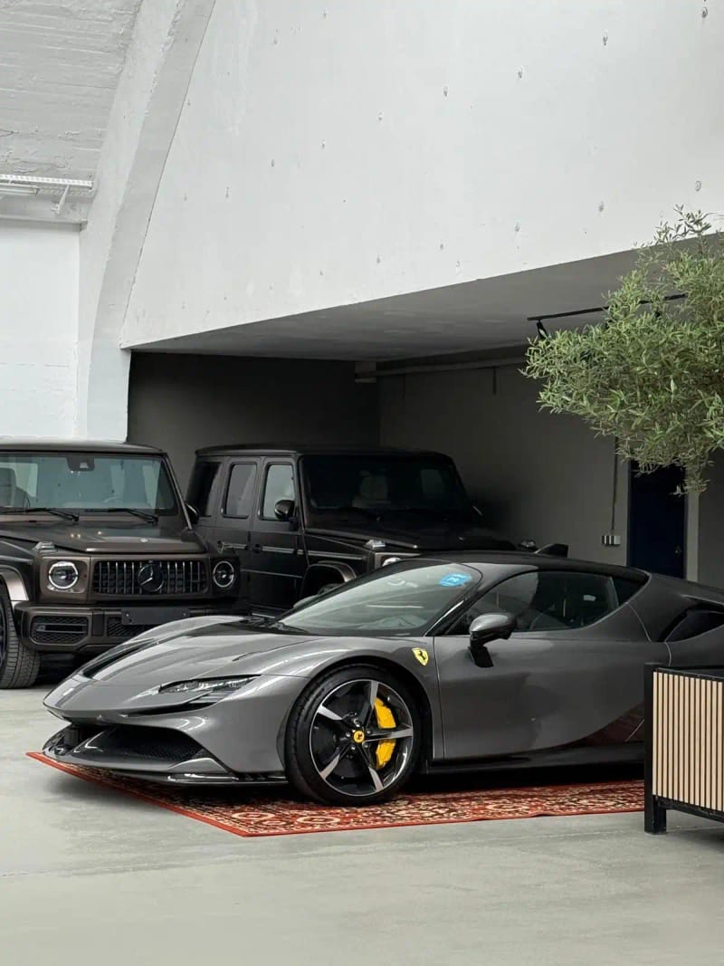Grauer Ferrari Sportwagen (seitliche Perspektive) mit gelben Bremssätteln auf orientalischem Teppich, geparkt in moderner Garage neben zwei schwarzen Mercedes‑G‑Klasse SUVs.